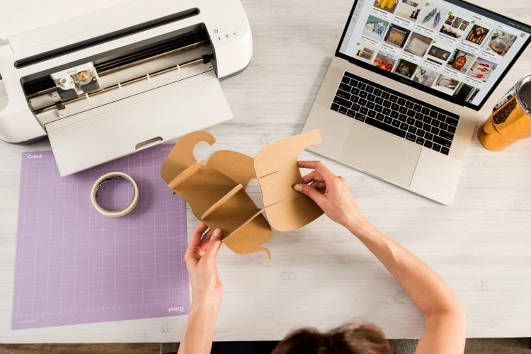 A laptop, a cricket machine, a roll of tape one desk with a person sitting at the desk assembling a cut out image