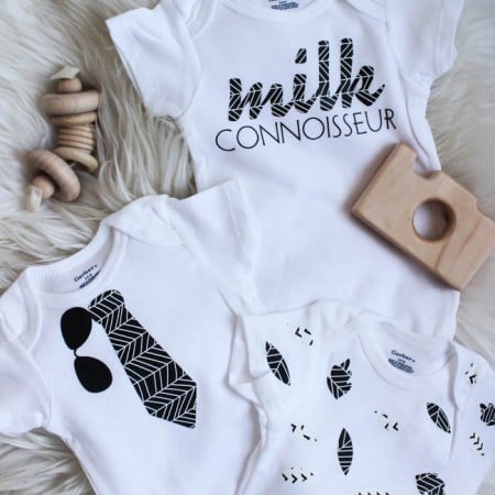 Some wooden baby toys laying on a furry rug along with three white onesies, one decorated with leaves, another one with a tie and sunglasses and the third one says, "Milk Connoisseur"