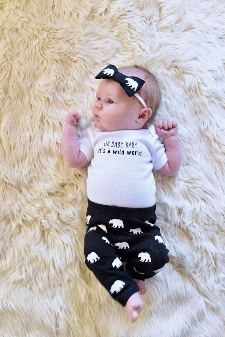 A baby lying on a furry rug, wearing a black pair of pants and a headband decorated with elephants and a white onesie that says, \"Oh Baby Baby It\'s a Wild World\"