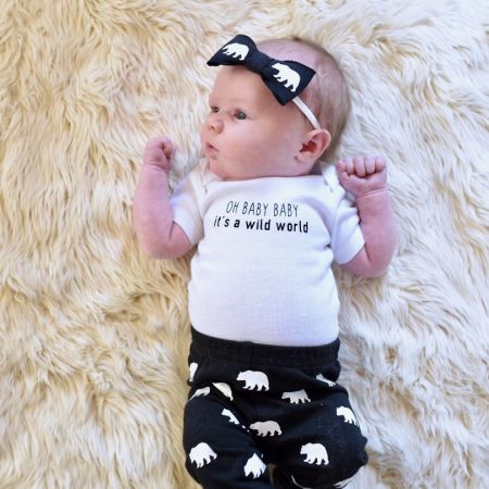 A baby lying on a furry rug, wearing a black pair of pants and a headband decorated with elephants and a white onesie that says, "Oh Baby Baby It's a Wild World"
