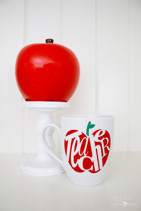 A red apple sitting on a pedestal next to a coffee mug that has an image of an apple and the word \"teacher\" on it
