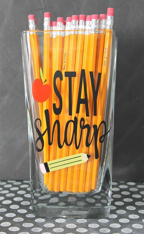 A clear water glass filled with pencils and decorated with an image of an apple and a pencil on it and the text, \"Stay Sharp\"