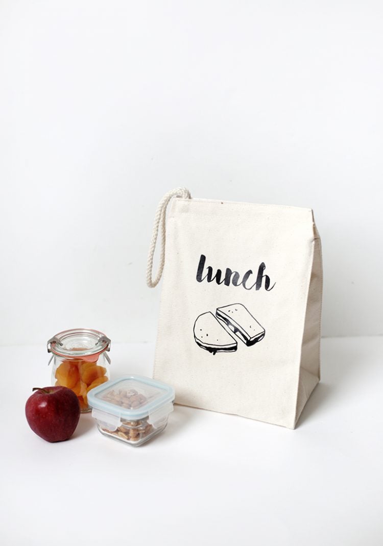 Two containers of food and an apple next to a canvas bag that has an image of a sandwich on it and the word \'lunch\'