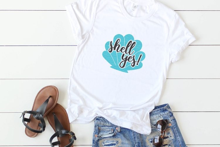 Close up of blue jean shorts, sandals and a white t-shirt decorated with a blue seashell and the saying, \"Shell Yes!\"
