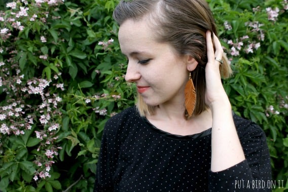A woman standing outside next to a floral bush pushing her hair back to show her earring