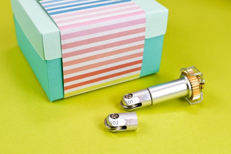 A close up of a Cricut scoring wheel tool sitting next to a box wrapped in multicolored striped paper and a paper rose on top of it
