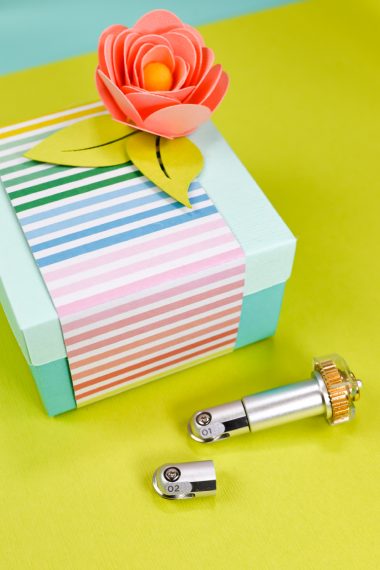 Cricut scoring wheel tool sitting next to a box wrapped in multicolored striped paper and a paper rose on top of it