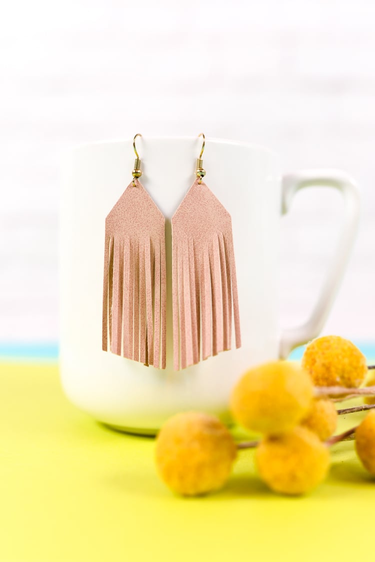 A pair of leather fringed earrings hanging from a coffee cup next to a bouquet of flowers
