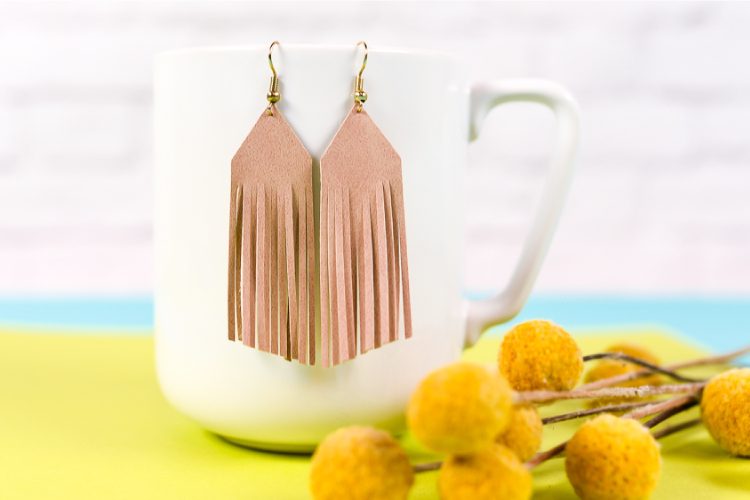 A pair of leather fringed earrings hanging from a coffee cup next to a bouquet of flowers