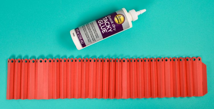 A bottle of tacky glue and accordion folded red paper that has holes punched in the top of each fold