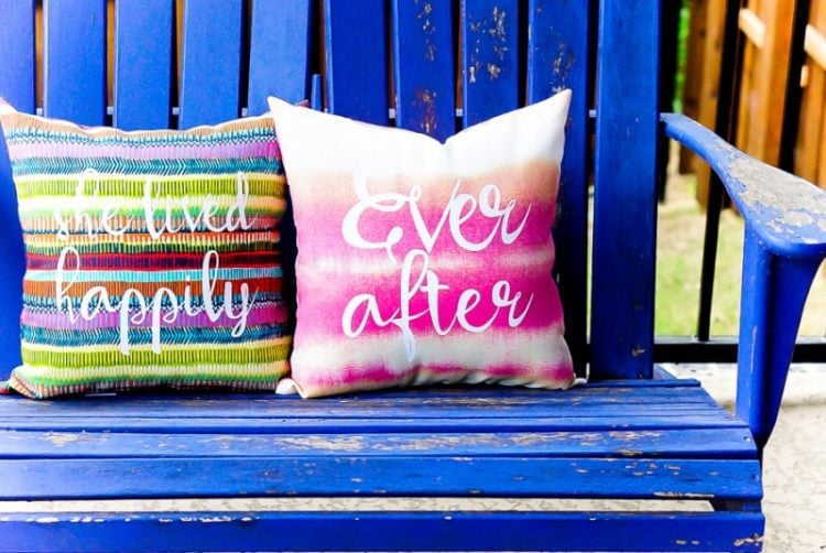 A blue bench sitting next to a wooden fence that contains two pillows, one says, \"She Live Happily\" and the other pillow says, \"Ever After\"