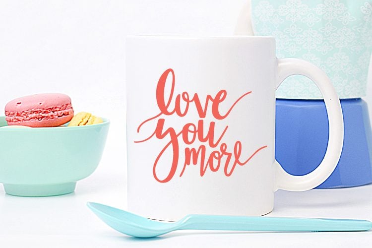 A canister sitting next to a spoon, a bowl of macarons and a coffee mug.  The mug is decorated with the text, \"Love You More\"