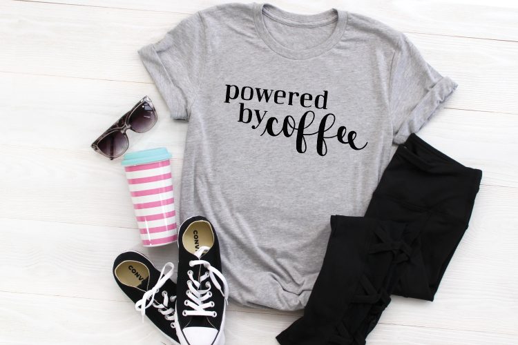 Close up of a pair of sunglasses, tennis shoes, black jeans, coffee tumbler and a gray t-shirt that says, \"Powered by Coffee\"