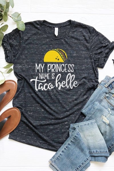 Some greenery, a pair of sandals, blue jeans and a gray shirt with an image of a taco on it and the saying, "My Princess Name is Taco Belle"