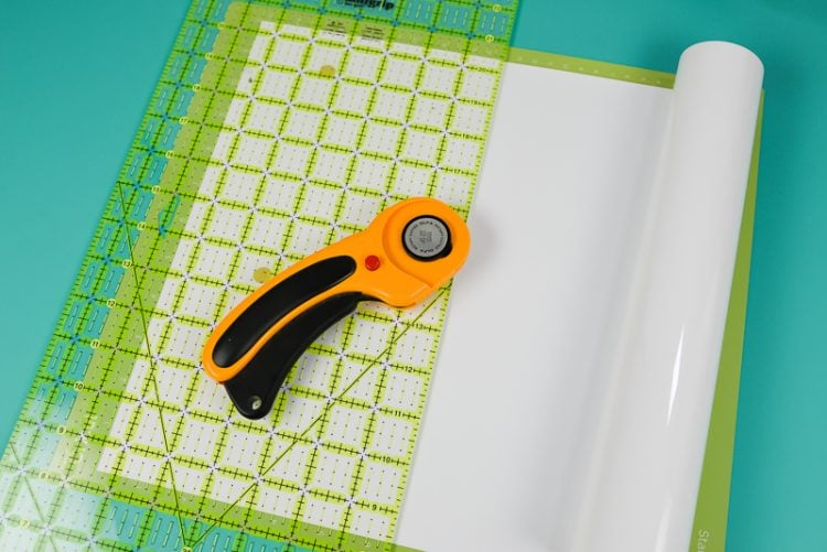 A rotary cutter and quilting ruler next to white vinyl adhered to a Cricut mat