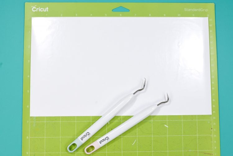 A green Cricut mat with white vinyl on it along with two weeding tools