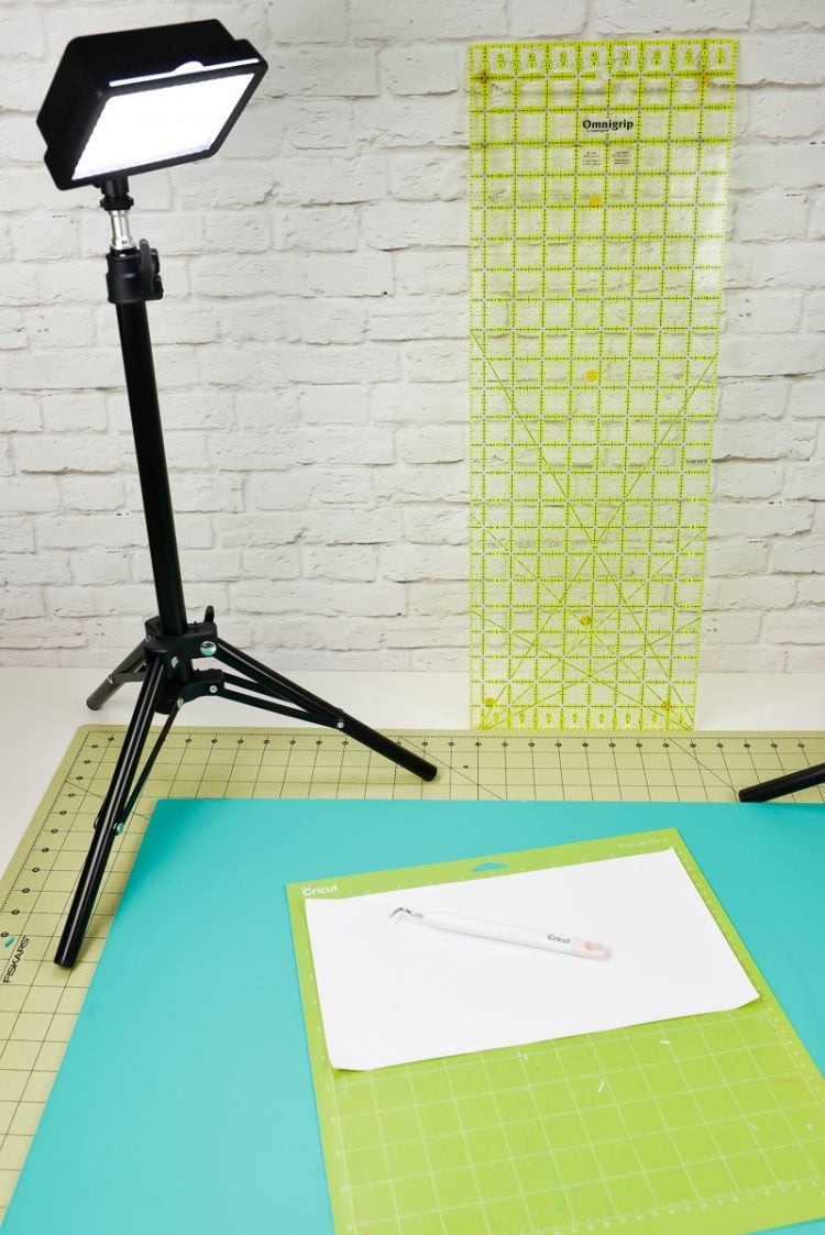 A studio photography light sitting on a desk that contains a green Cricut mat that has white vinyl and a weeding tool on top of it