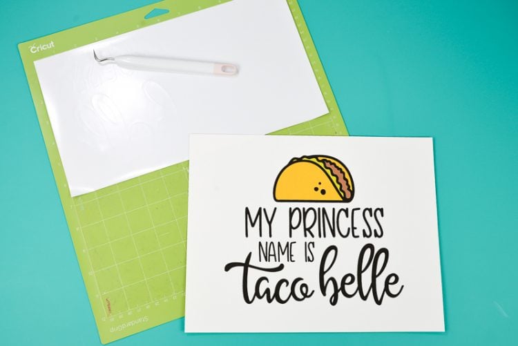 A weeding hook, on top of a piece of white vinyl that is on a Crciut mat and a piece of paper with a design printed on it of a taco and the text, \"My Princess Name is Taco Belle\"