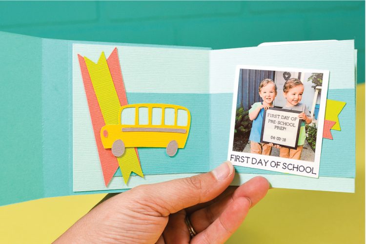 A hand holding a back-to-school memory book with an image of a school bus cut out of paper and a picture of two adorable little boys holding a sign that says, \"First Day of Pre-School Prep\"