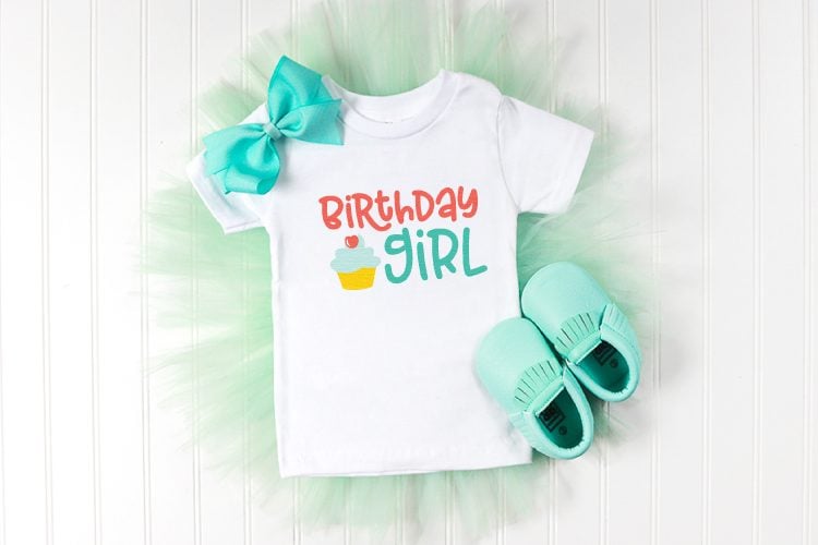 Little aqua colored moccasins, an aqua colored hair bow, and a white t-shirt decorated with an image of a cupcake and the text, \"Birthday Girl\"