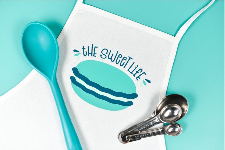 Close up of measuring spoons, large mixing spoon and an apron with the saying, \"The Sweet Life\" and an image of a macaron