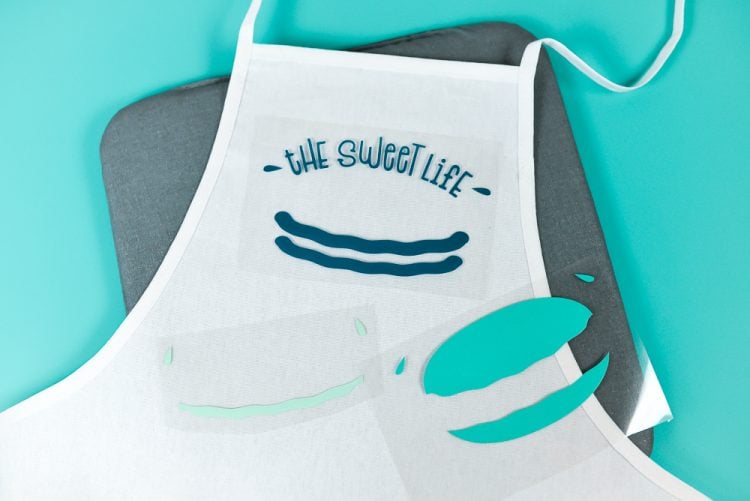 An apron lying on top of a mat, with the saying, \"The Sweet Life\" and the vinyl pieces that make up a macaron yet to be applied