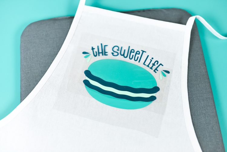 An apron lying on top of a mat, with the saying, \"The Sweet Life\" and an image of a macaron on it 