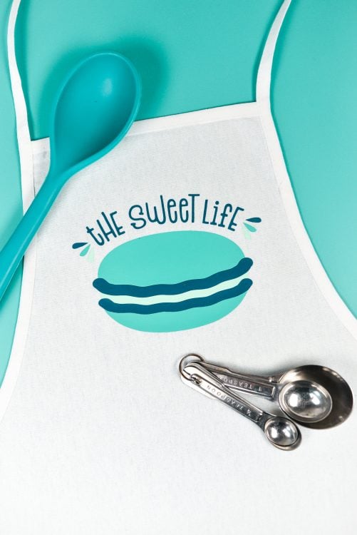 Close up of measuring spoons, large mixing spoon and an apron with the saying, "The Sweet Life" and an image of a macaron
