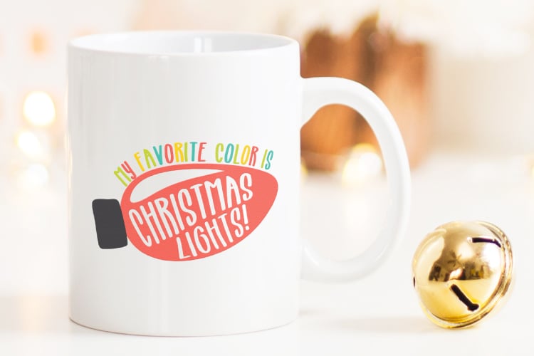 A close up of a jingle bell and a coffee mug on a table, with and image of a Christmas light with the saying, \"My Favorite Color is Christmas Lights!\"