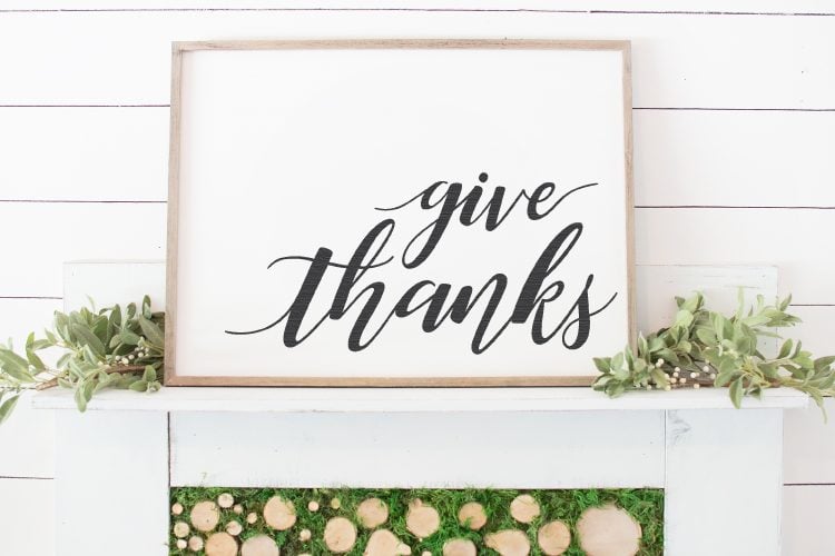 A close up of a wood framed white sign that says, \"Give Thanks\" and is surrounded with decorative greenery