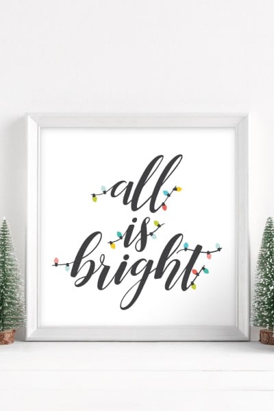 Two small trees next to a white framed sign that says, "All Is Bright"