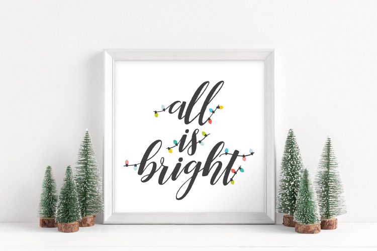 Six small trees next to a white framed sign that says, \"All Is Bright\"