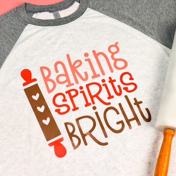 Close of a rolling pin and a baseball style shirt with gray sleeves and white body that has an image of a rolling pin and saying, \"Baking Spirits Bright\"