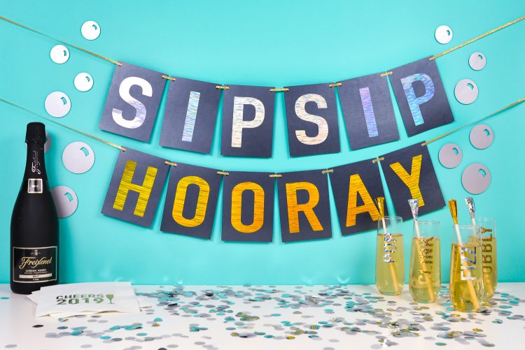 A bottle of champagne, some 2019 napkins, glasses of champagne all sitting under a banner that says, \"Sip Sip Hooray\"