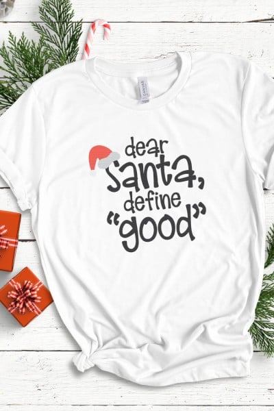 A white shirt lying next to Christmas decor and decorated with a design that says, "Dear Santa, Define "Good'"
