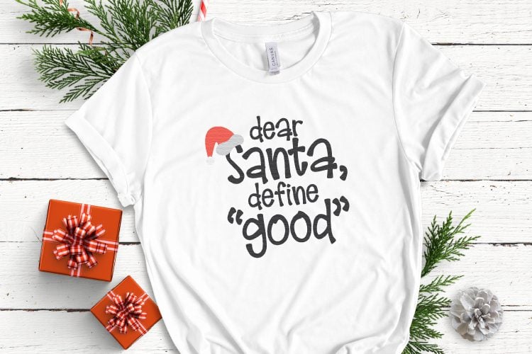 A close up of a white shirt lying next to Christmas decor and decorated with a design that says, \"Dear Santa, Define \"Good\'\"