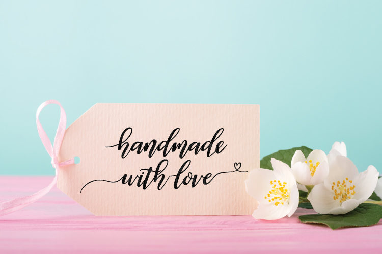 Close up of flowers on a pink table lying next to a tag with a pink bow tied to it and the lettering \"Handmade with Love\" on it