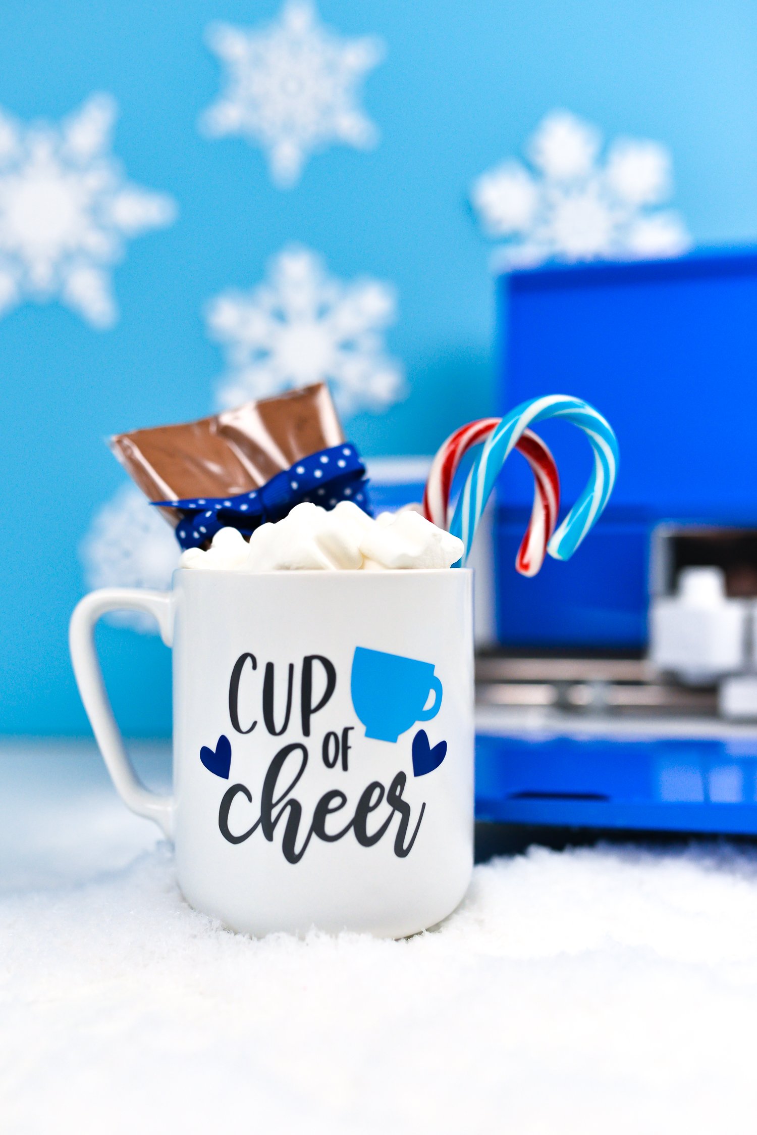 Close up of a Cricut machine in the background with a mug filled with candy canes, marshmallows and package of hot cocoa mix and decorated with an image that contains a coffee cup and hearts and says, \"Cup of Cheer\"