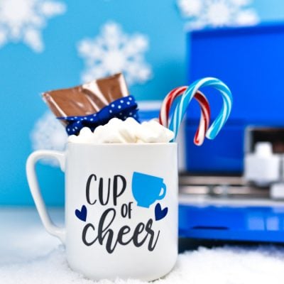 A Cricut machine in the background with a mug filled with candy canes, marshmallows and package of hot cocoa mix and decorated with an image that contains a coffee cup and hearts and says, "Cup of Cheer"