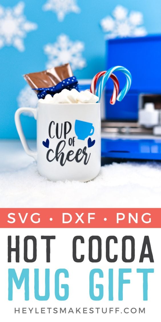A Cricut machine in the background with a mug filled with candy canes, marshmallows and package of hot cocoa mix and decorated with an image that contains a coffee cup and hearts and says, \"Cup of Cheer\"
with advertising a Hot Cocoa Mug Gift idea from HEYLETSMAKESTUFF.COM