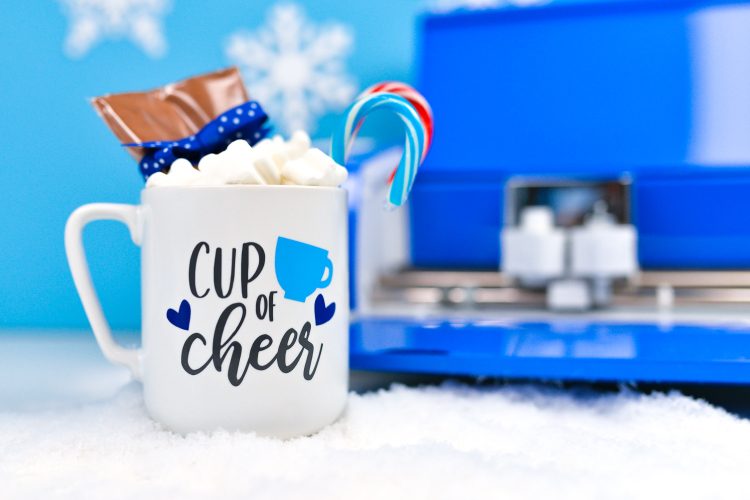 Close up of a Cricut machine in the background with a mug filled with candy canes, marshmallows and package of hot cocoa mix and decorated with an image that contains a coffee cup and hearts and says, "Cup of Cheer"