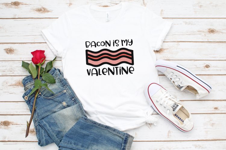 A pair of blue jeans, a long-stemmed red rose, tennis shoes and a white t-shirt that is designed with a piece of bacon and says, \"Bacon is my Valentine\"