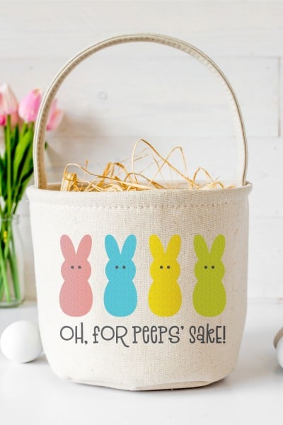 A vase of pink tulips and some undyed eggs next to a canvas style Easter basket that is decorated with 4 Easter bunnies and says, "Oh, for Peep's Sake!"