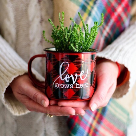 Woman holding a red and black buffalo patterned coffee mug that has a plant in it and the saying Love Grows Here