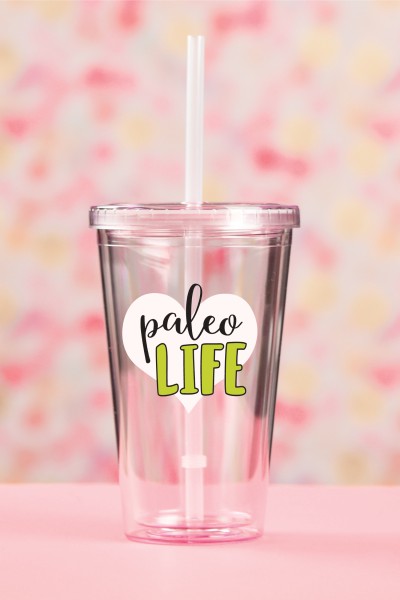 A pink table against a pink and yellow blurred background holding a clear plastic glass with a straw in it with a design on it that says, "Paleo Life"