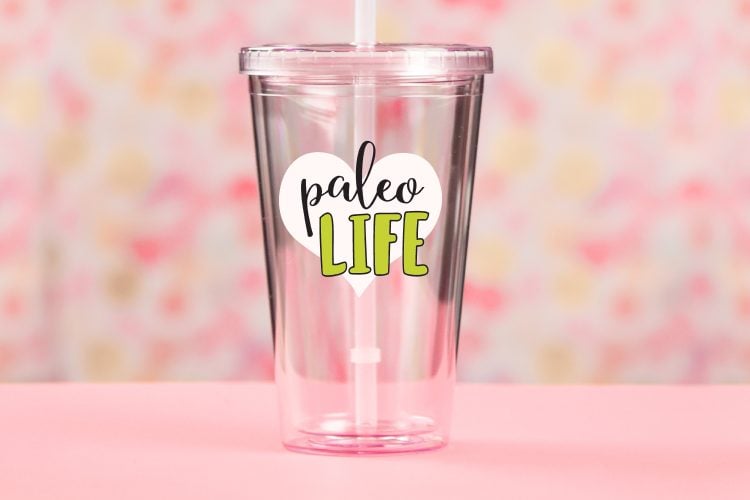 A pink table against a pink and yellow blurred background holding a clear plastic glass with a straw in it with a design on it that says, \"Paleo Life\"