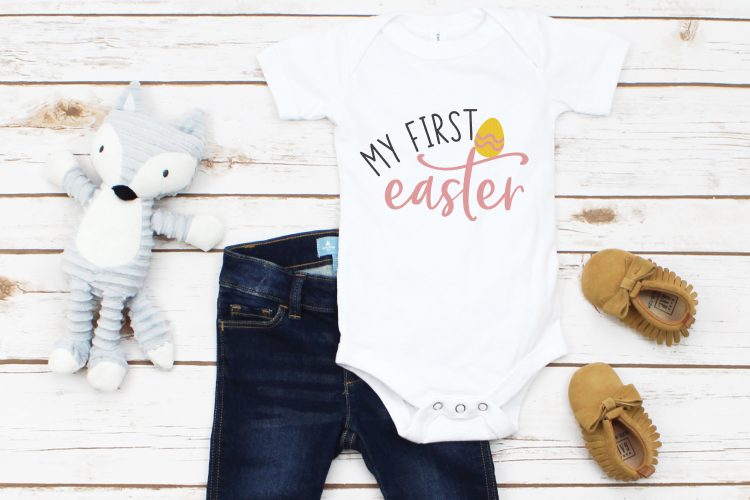 A little gray stuffed toy fox, a pair of baby blue jeans, moccasins and a white onesie decorated with an Easter egg and the saying, \"My First Easter\"