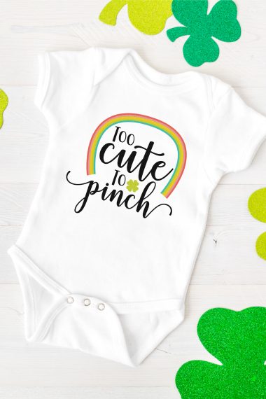 Green shamrocks around a white onesie that is decorated with a rainbow and the saying, "Too Cute to Pinch"