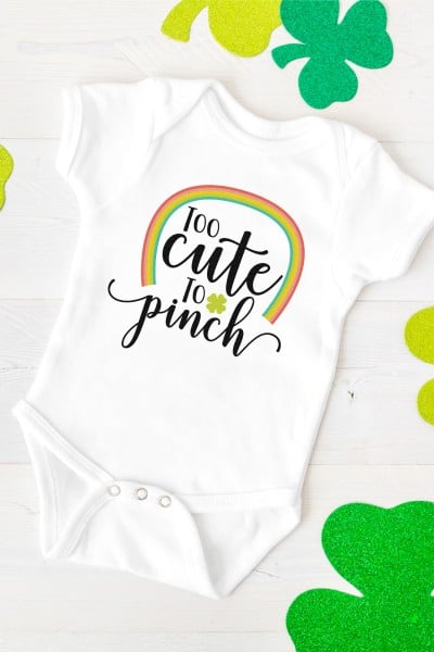Green shamrocks around a white onesie that is decorated with a rainbow and the saying, "Too Cute to Pinch"