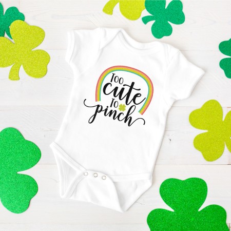 Close up of green shamrocks around a white onesie that is decorated with a rainbow and the saying, "Too Cute to Pinch"
